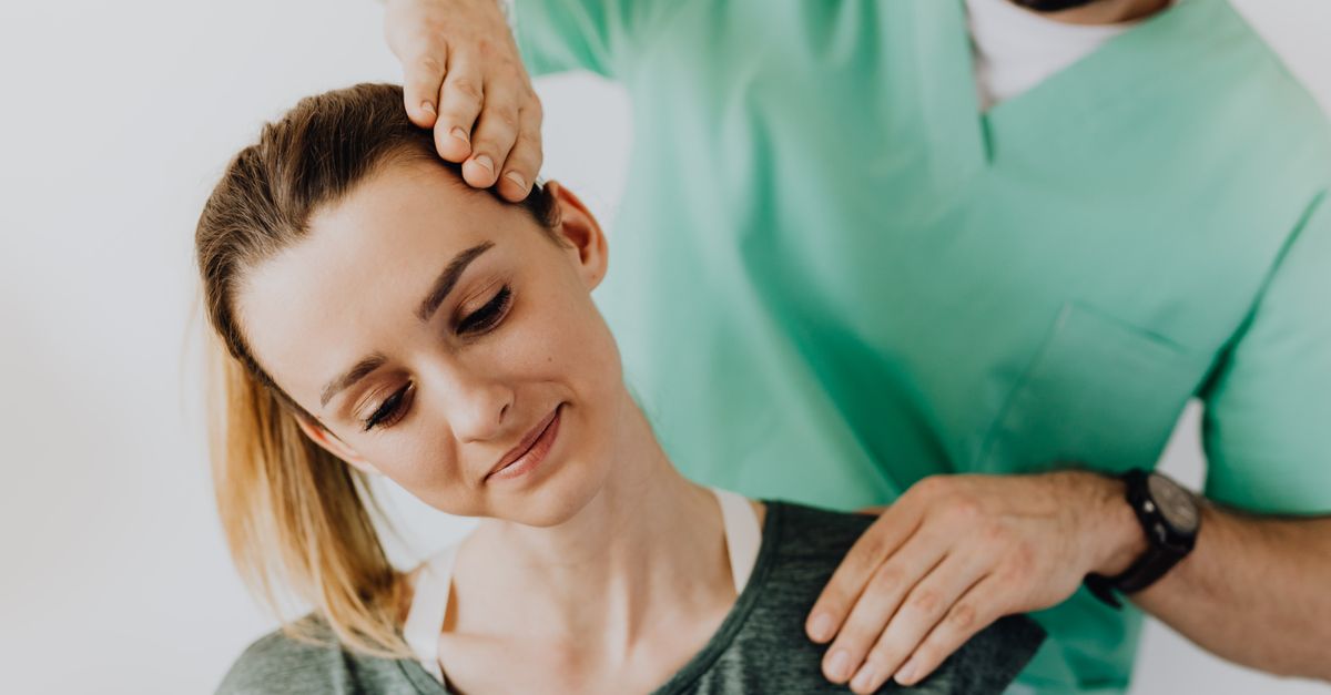 Serious doctor in uniform massaging smiling female patients neck and stretching stiff neck muscles against gray background