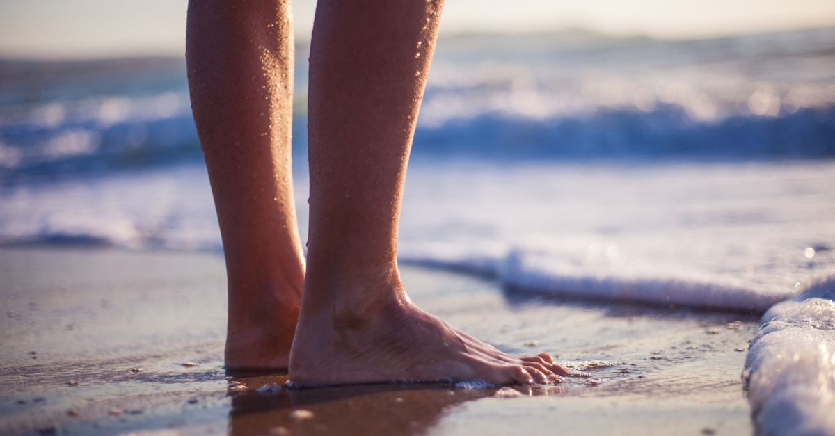Close-up of barefoot person standing on the beach with ocean waves.