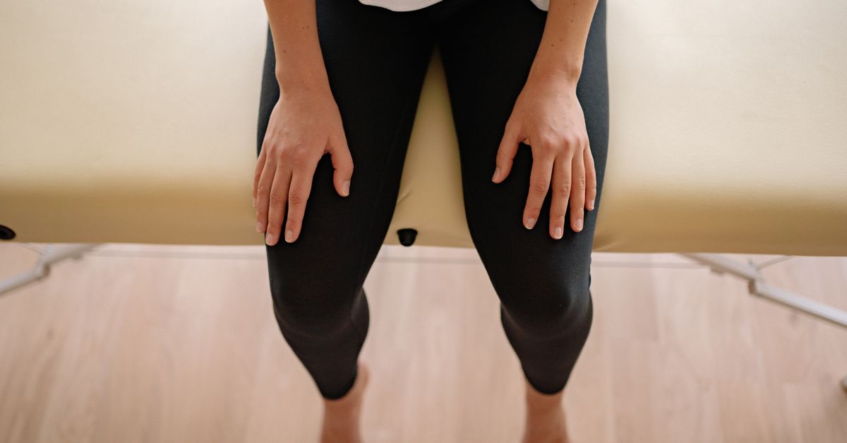 A woman sitting barefoot on a massage table indoors, captured from above.