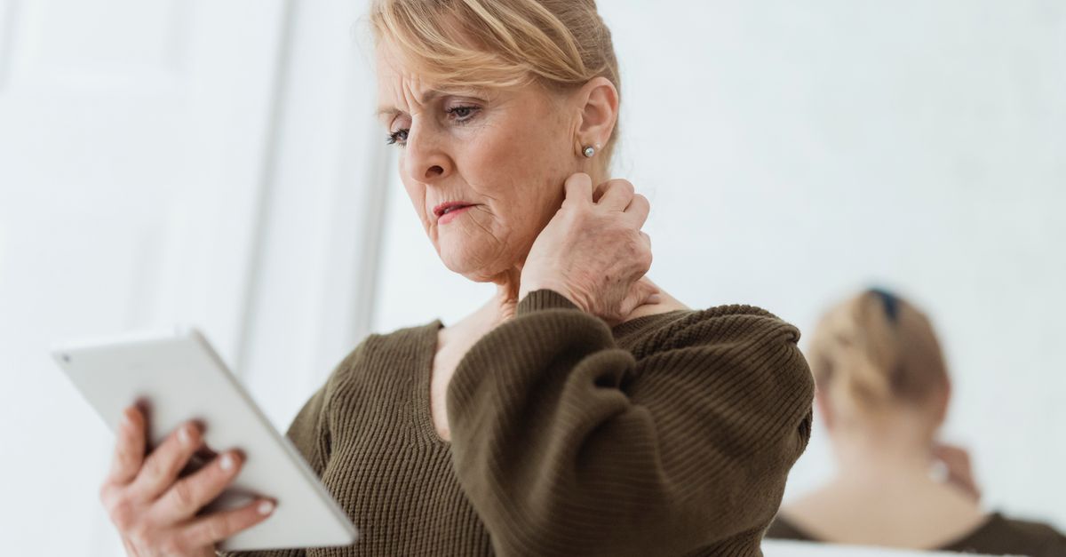 A contemplative mature woman in casual wear touching her neck in a light room.