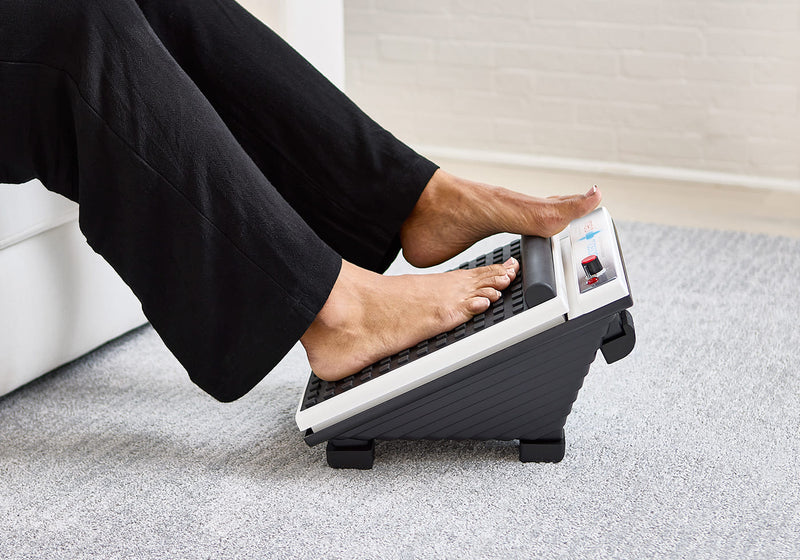 Person uses a black foot massager on a light carpet, sitting with both feet placed on the device.