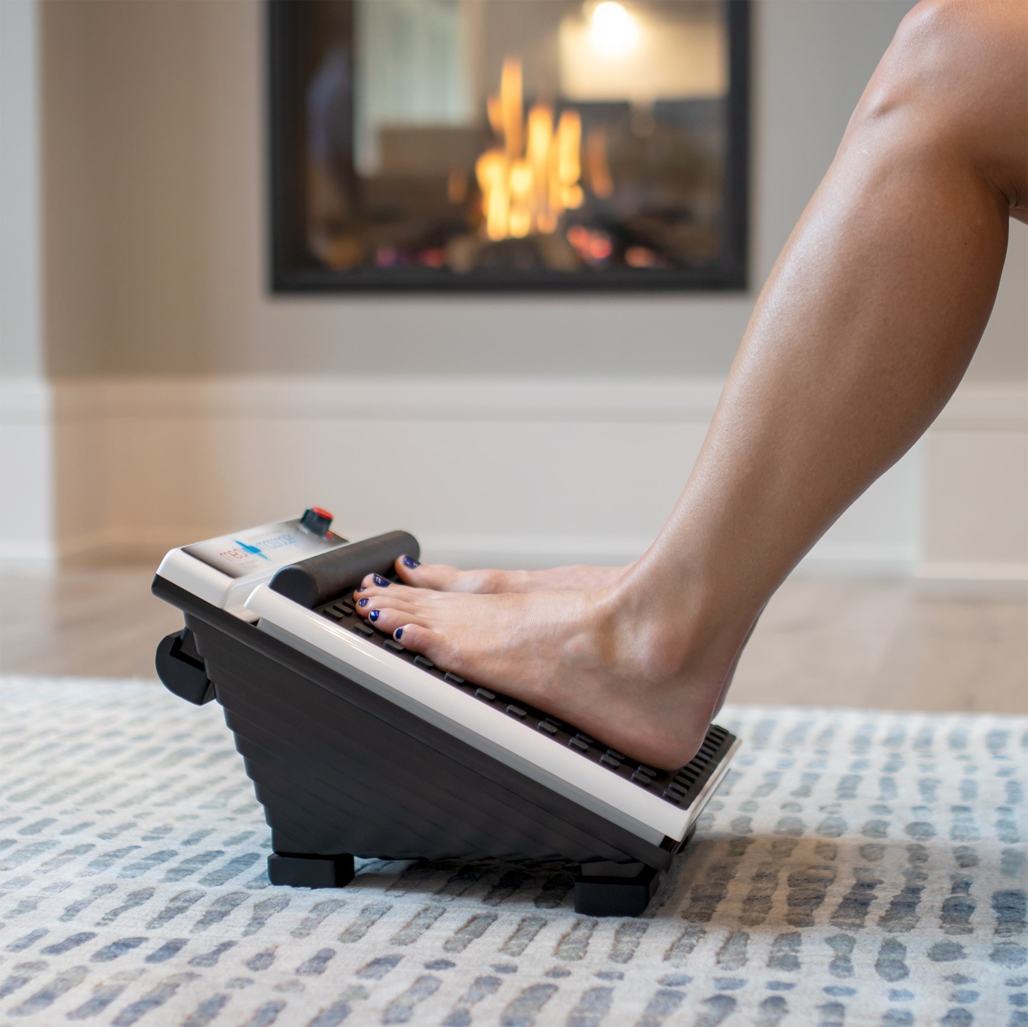Person using MedMassager foot massager on a rug by the fireplace for at-home relaxation and relief.