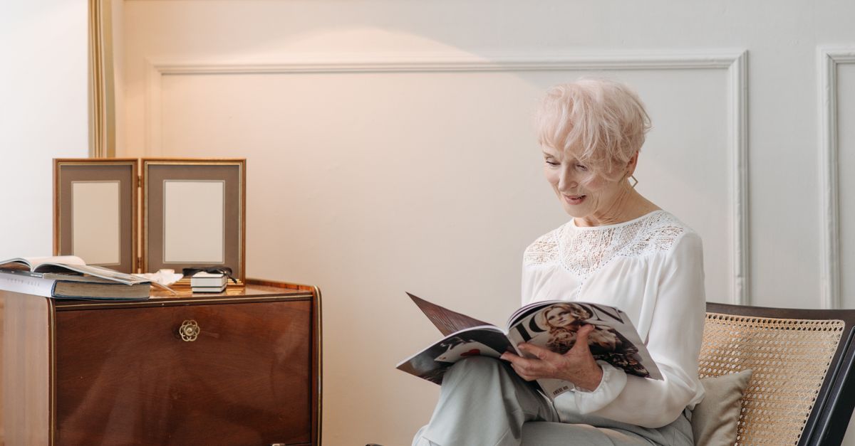 Elderly woman reading a magazine on a comfortable chair in a well-lit living room.