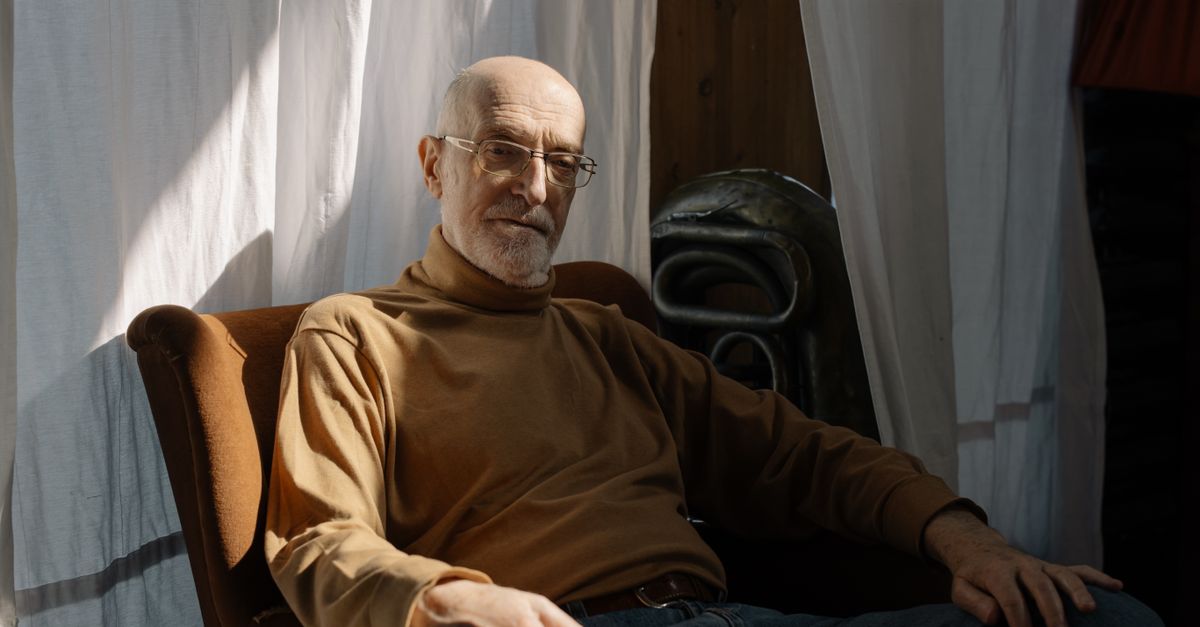 Elderly man with glasses sitting in an armchair in a warmly lit room.