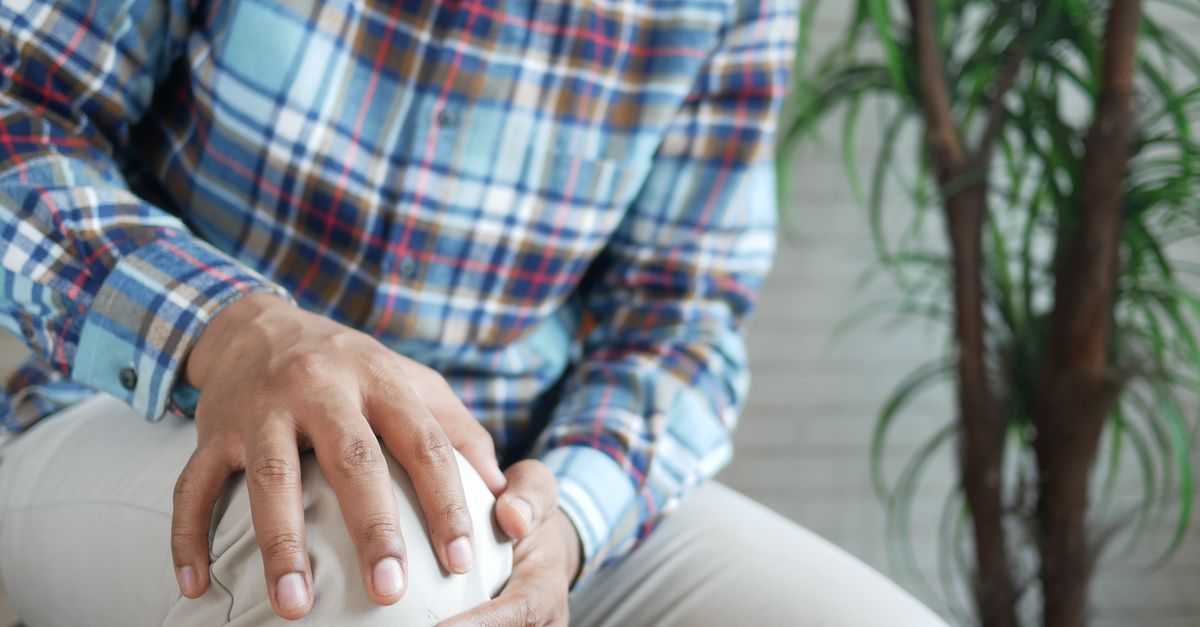 Close-up of a man indoors in a plaid shirt, holding his leg, indicating stiffness or discomfort.