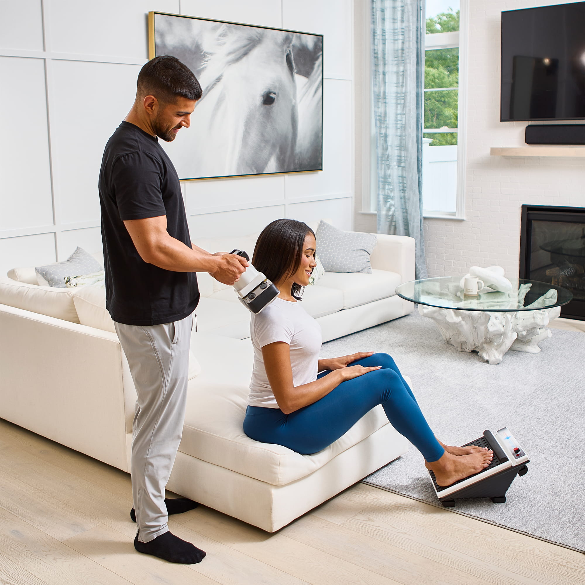 Man uses MedMassager foot massager while woman relaxes in bright living room.