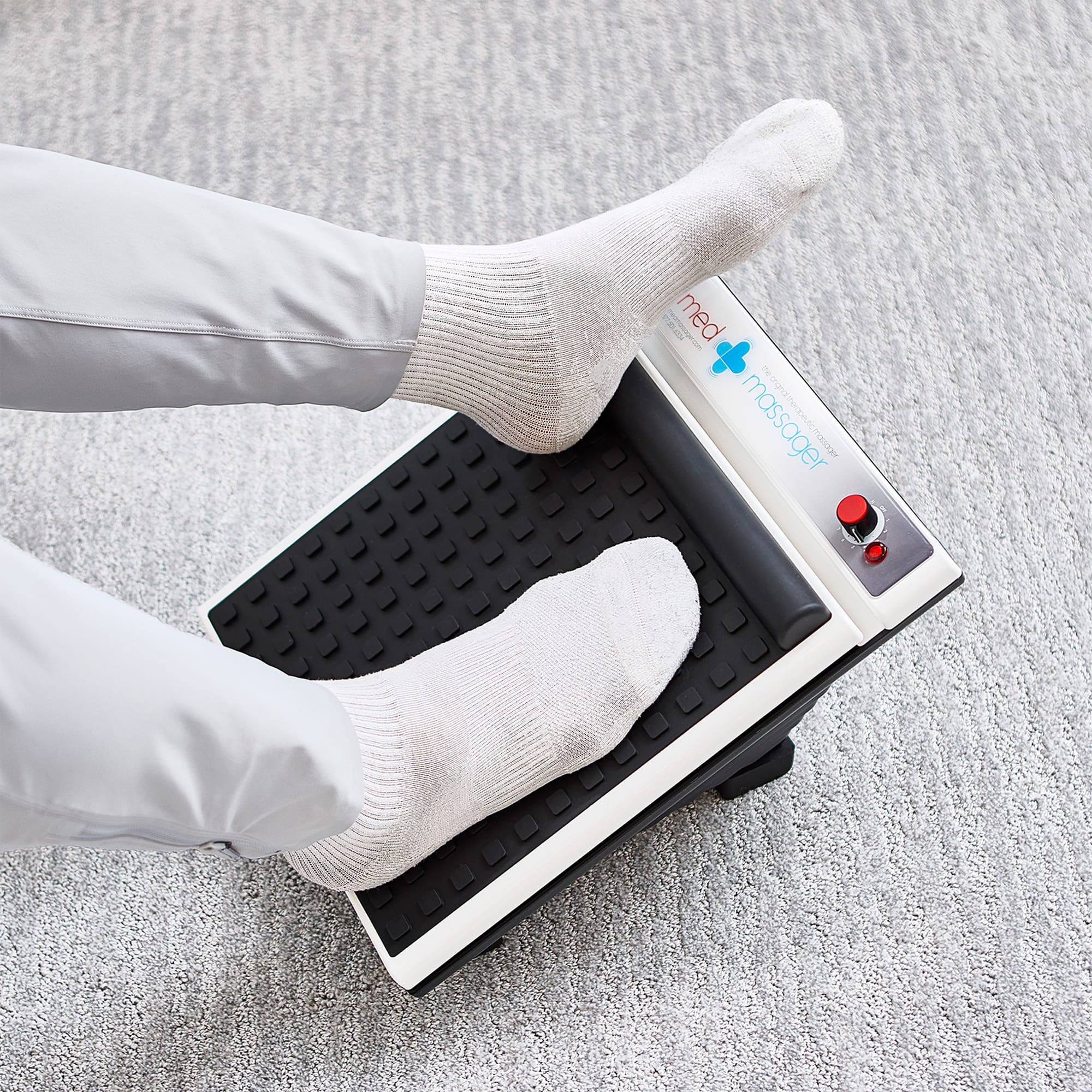 Person in white socks using MedMassager foot massager on a light carpet.